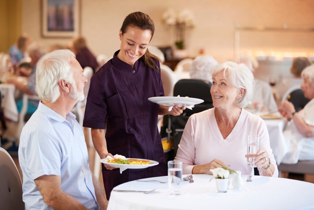 Senior couple being served a meal by resident staff in the dining room of retirement home