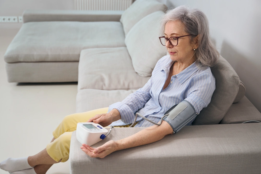 Elderly woman sits on a sofa at home and measures blood pressure with a tonometer
