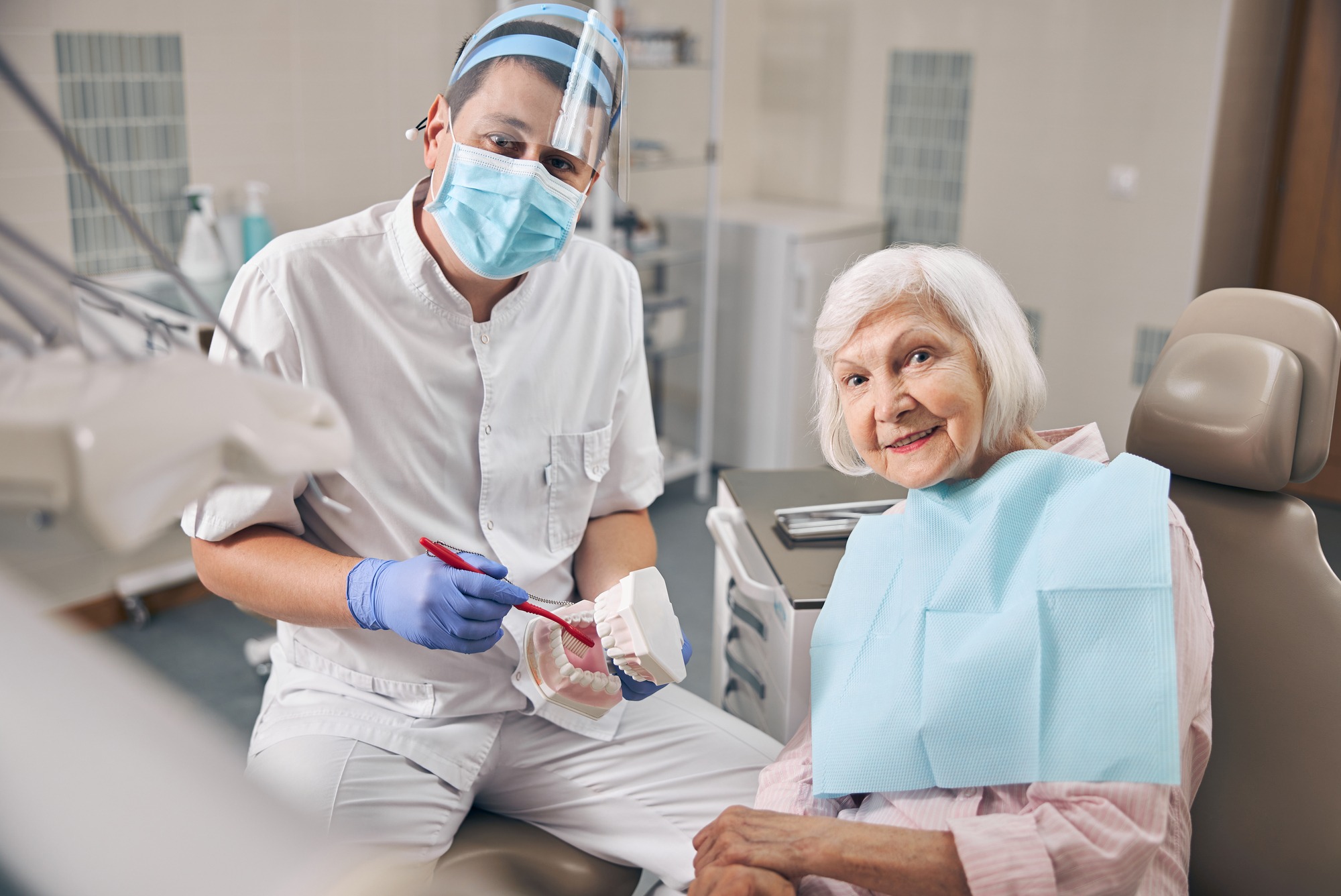 A young doctor in uniform is telling a senior woman about teeth in a dental office