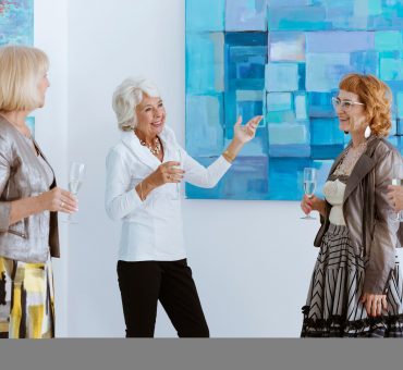 Group of senior women smiling and drinking sparkling wine as they tour an art gallery