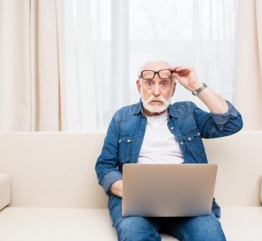Shocked senior man sitting with laptop on knees and holding eyeglasses