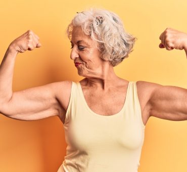 Senior grey-haired woman wearing casual clothes showing arms muscles smiling proud.